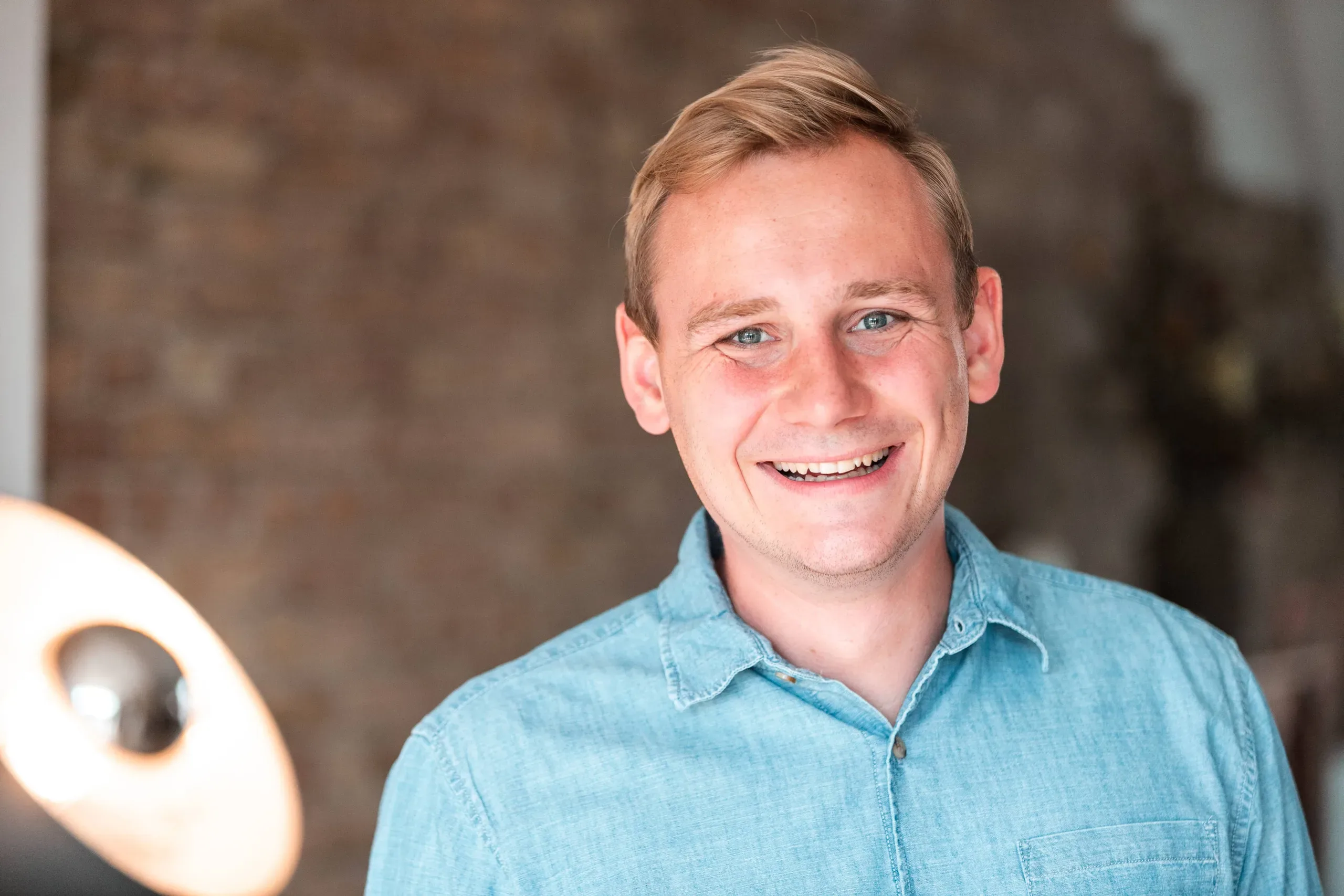 A blond man in a blue denim shirt smiles in front of a brick wall with a lamp to his side.