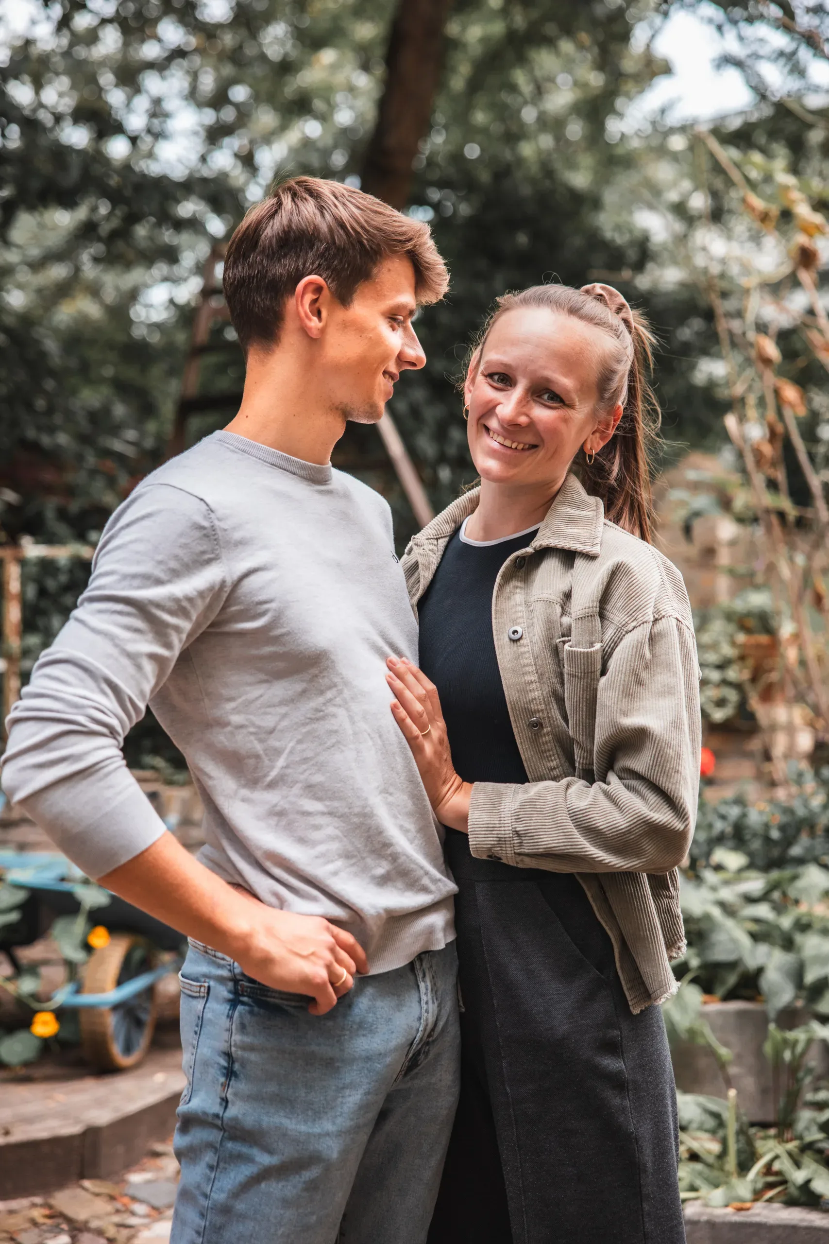 A man in a grey long-sleeved shirt and a woman in a beige corduroy jacket stand close together in a garden, with the woman smiling toward the camera and resting her hand on his chest.